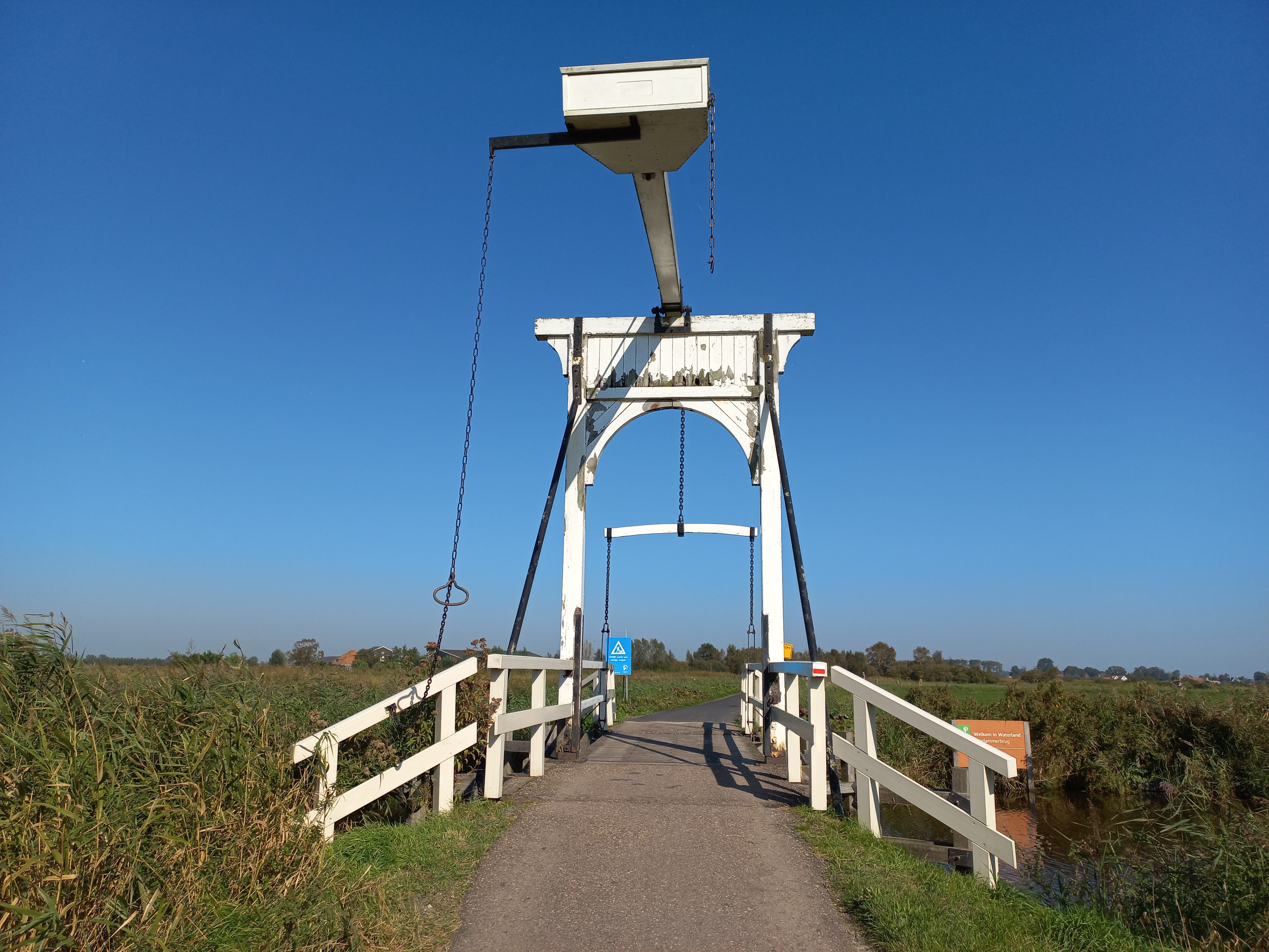 The white timber Aandammerbrug lift bridge spans a narrow reed-lined waterway beneath a clear blue sky.