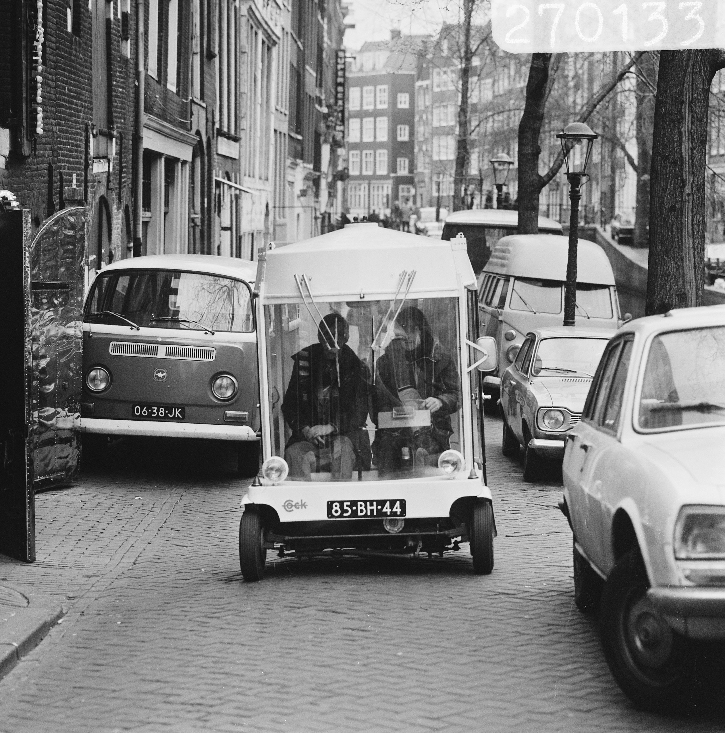 A small white cylindrical Witkar carries two people along a crowded Amsterdam street in 1974.